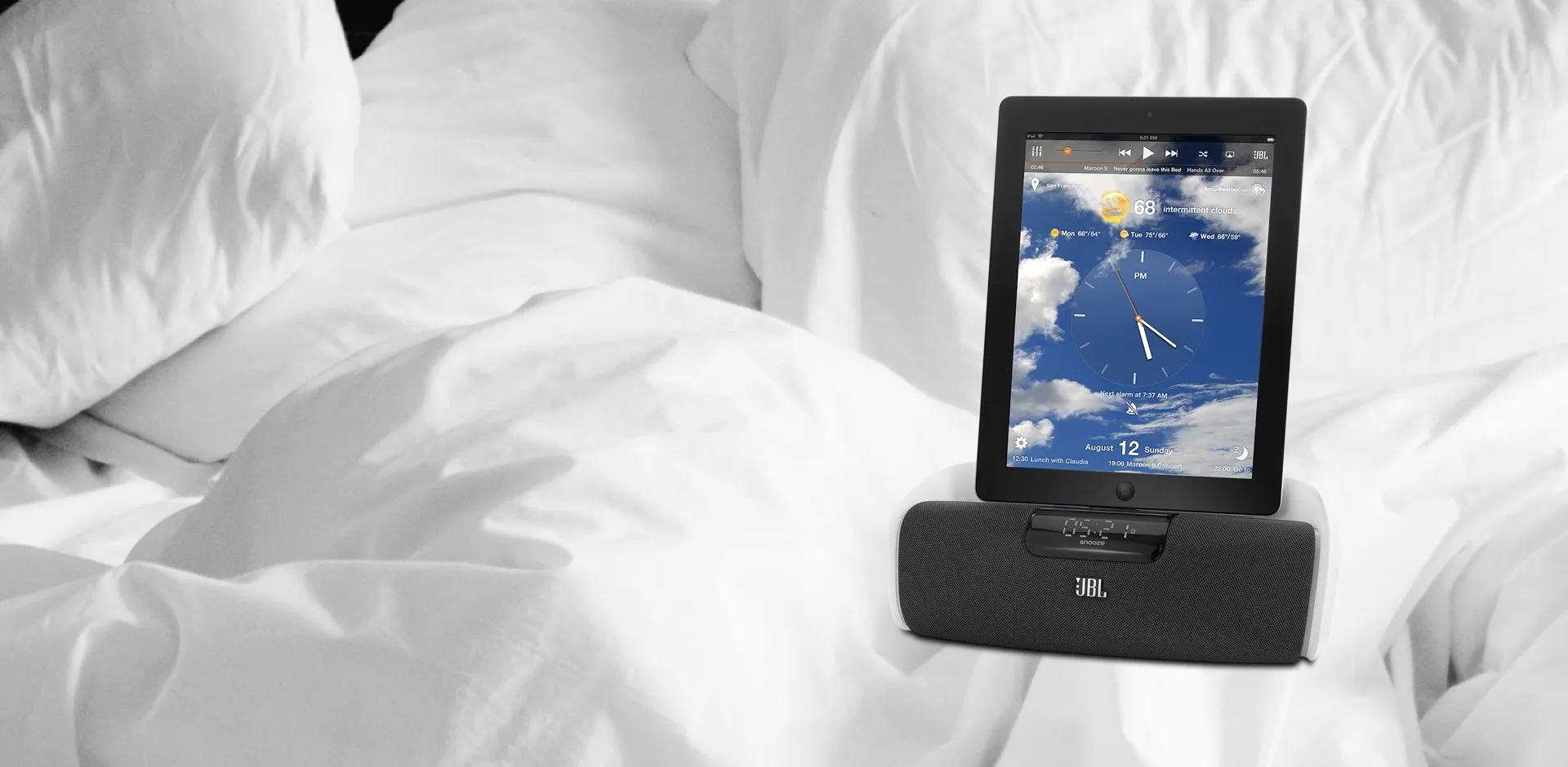 A tablet docked on a JBL brand speaker dock shows an interface of a music player, large clock, and weather background.