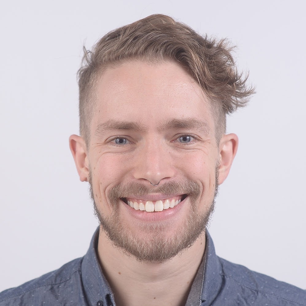 Headshot of a smiling person wearing a blue shirt, with waved hair parted to the side, and a light beard.